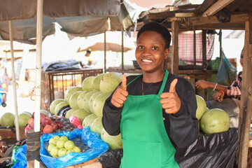African woman or female trader with a green apron, doing thumbs up gesture while standing at her...