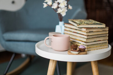 Pink cup of tea, chocolate pralines and books on a small table, decorated with flowers, armchair in background, copy space