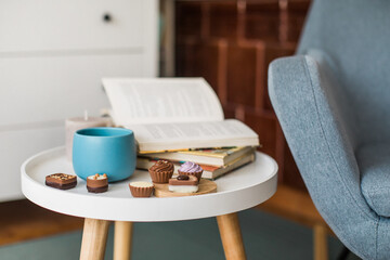 Book, cup of coffee and chocolate pralines on round white side table in living room, Scandinavian...