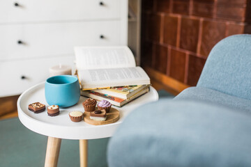 Side view photo of book, cup of green tea and chocolate pralines on round white side table, Scandinavian design, copy space