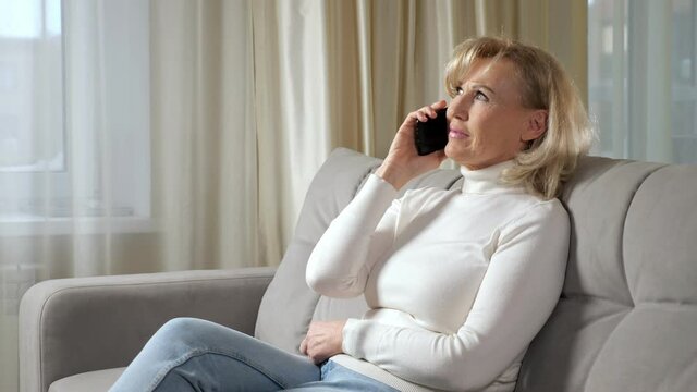 Blonde-haired woman of mature age talks via smartphone and smiles broadly relaxing on comfortable sofa in living room against window closeup.