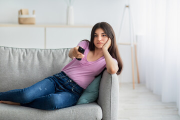 Bored Indian teen girl with remote control watching TV on sofa at home, copy space