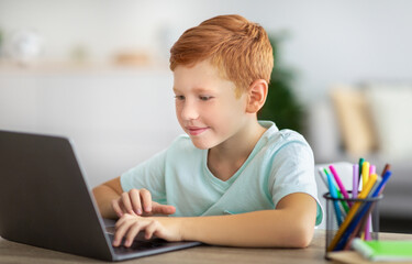 Joyful kid ginger boy doing homework, using laptop