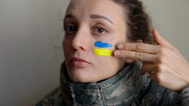Young girl is paining blue and yellow ukrainian flag on her cheek using her fingers wearing military uniform, mandatory conscription in Ukraine, equality concepts
