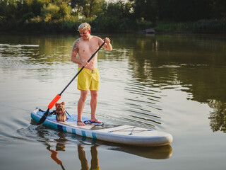 Young person stand on paddle board with his small yorkshire terrier and swimming on a river in a sunny day. Stand up paddle boarding or paddling is awesome active recreation in nature.