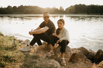A young father with beard is sitting with his daughter on the coast of the lake