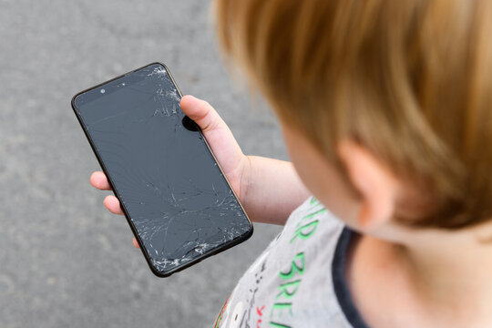 The Boy Holds In His Hand A Smartphone With A Broken Screen On The Background Of Asphalt. A Child Dropped An Expensive Phone On The Asphalt.