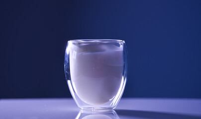 At transparent glass filled with froth milk, double wall thermo resistant glass, standing on a white table with dark background.