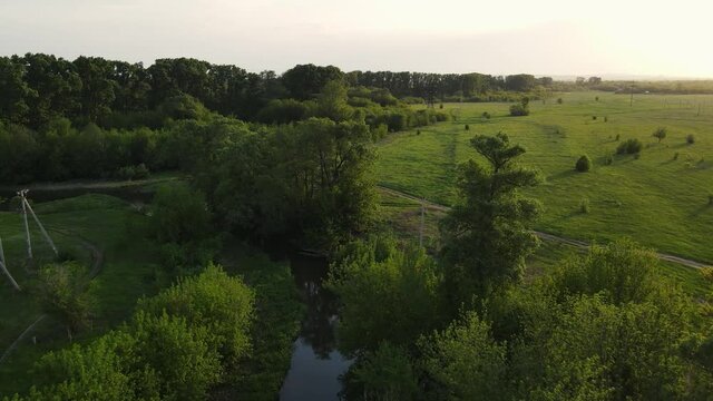 4k footage from a drone moving along a green field with a river and a forest