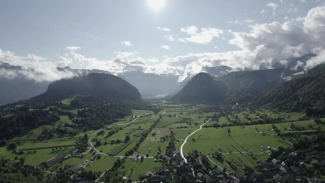 Reverse plane drone video over farms and houses in the Triglav with hills on the horizon, the sun in front and clear sky with clouds.
En Eslovenia, Bohinj.