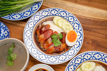 Rice with red roasted pork, Crispy Pork with sauce and Egg topped in a white dish on wooden table.