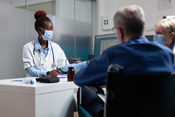 Fototapeta premium Physician doctor wearing medical face mask to prevent infection with coronavirus in hospital office. Therapist woman explaining disease diagnosis to elderly people discussing healthcare treatment