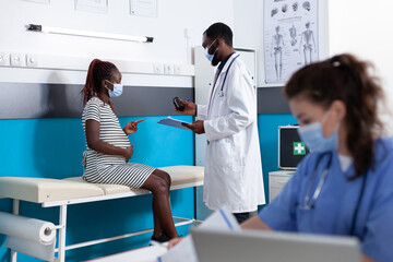 Obraz premium Doctor giving bottle of pills to pregnant woman at consultation. Pregnant patient talking to specialist at checkup visit, expecting child during pandemic. People wearing face masks