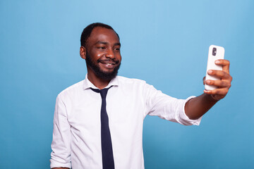 Smiling businessman having fun taking a selfie photo using smart phone front camera over blue background. Happy freelancer holding smartphone in video call conference posing casual.