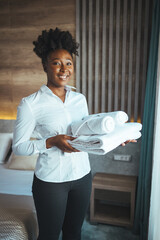 Hotel staff setting up pillow on bed. African housekeeper looking at the camera holding a towel a...