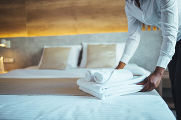 Chambermaid in uniform holding stack of clean folded towels while going to put them on changed bed in hotel room. Maid with fresh clean towels during housekeeping in a hotel room