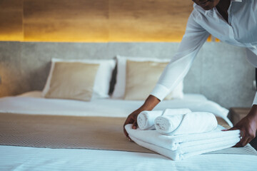 Young hotel maid making the bed with clean fresh towels. Hands of hotel maid bringing fresh towels to the room. Close up of a young hotel maid holding clean folded towels - obrazy, fototapety, plakaty