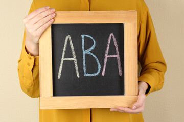 Woman holding small chalkboard with abbreviation ABA (Applied behavior analysis) on beige background, closeup - obrazy, fototapety, plakaty