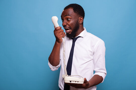 Angry Businessman Shouting Into Reciever Of Cord Telephone Arguing To Person Listening At The Other End Of The Line. Stressed Employee Having A Fight With Manager Over The Phone.