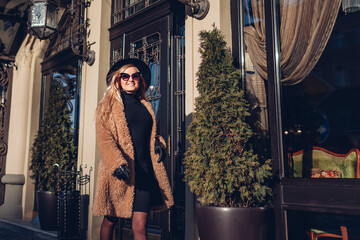 Stylish young woman walking in teddy coat hat sunglasses on city street outdoors. Spring fashionable female accessories.