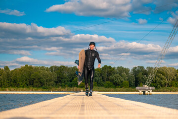 Athletic man in wetsuit carrying wakeboard, walking on pier after training