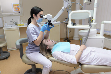 Young female dentist treats root canals using a microscope in a dental clinic. A female patient lies on a dentist chair with her mouth open.