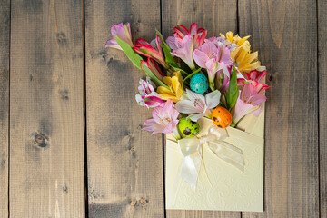 colorful eggs and blooming alstroemeria flowers in festive envelope with bow on wooden table. concept of easter, summer, spring, nature bloom, holiday and congratulations.copy space.flat lay.top view