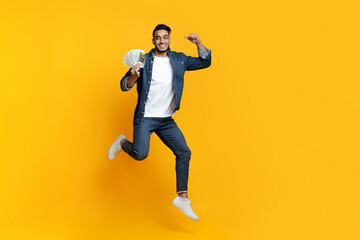 Positive middle-eastern man holding money on yellow background, celebrating win