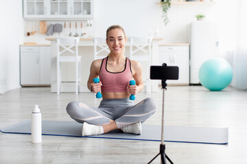 Cheerful european millennial blonde female fitness blogger look at webcam and make video of training with dumbbells
