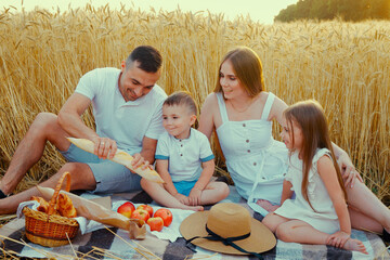 Happy family in summer outfits sitting on blanket and having picnic in field. Father dividing...