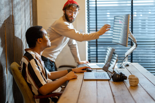 Two men with different nationality working on computers, sitting together at cozy home office. Concept of freelance and remote work. Stylish male hipsters programming together - Powered by Adobe