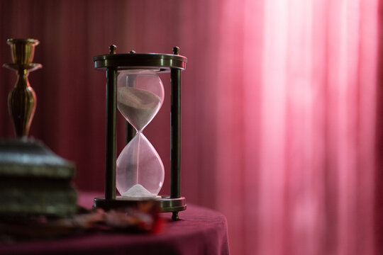 Photo of an hourglass on a table against the backdrop of a red curtain. Interior and time concept