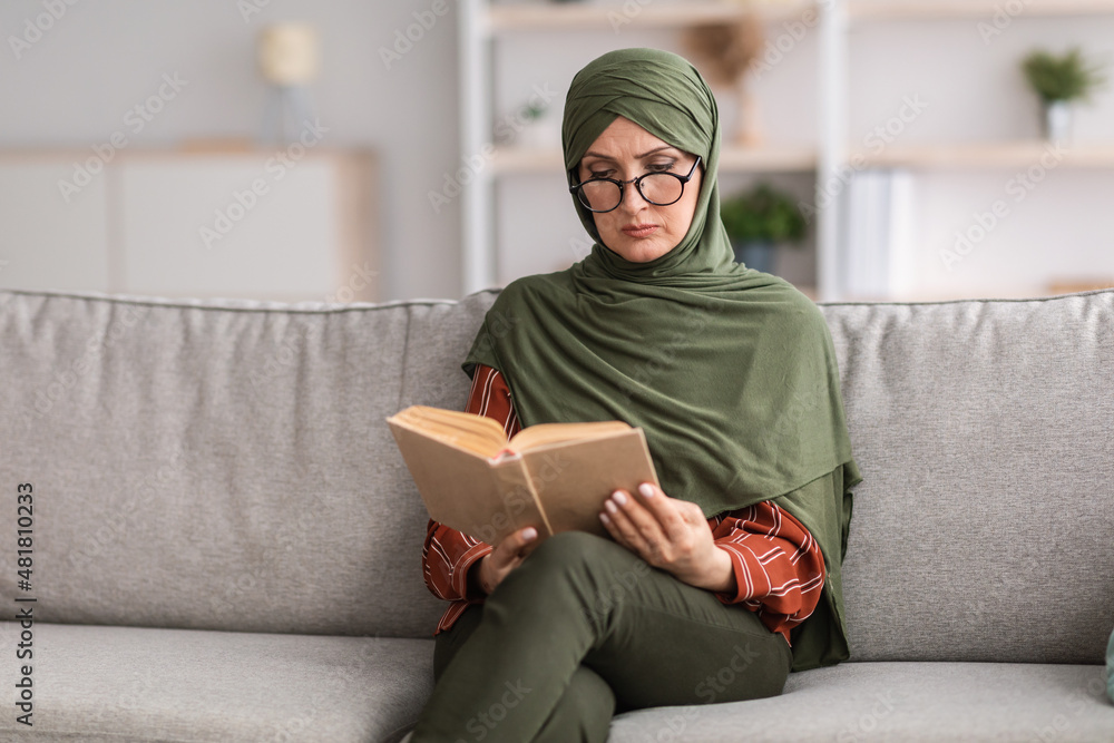 Wall mural Mature Muslim Female Reading Book Wearing Eyeglasses Sitting At Home