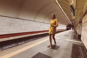 Walking on the subway platform beautiful young girl with long legs in a yellow spring coat and a white handbag in her hand.