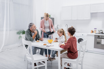 middle aged woman with frying pan near husband and grandchildren having breakfast in kitchen.