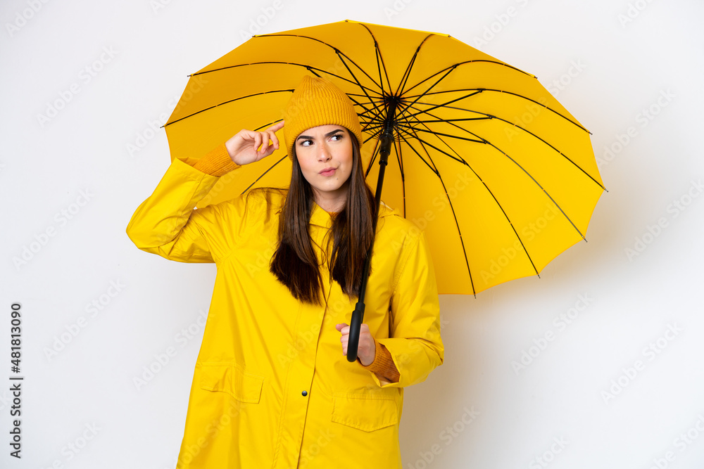 Wall mural Young Brazilian woman with rainproof coat and umbrella isolated on white background having doubts and thinking