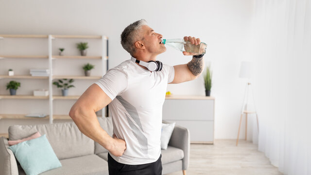 Stay hydrated. Mature man drinking water during home workout, standing in living room with headphones on neck