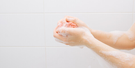 A girl in a white bathroom in foam rubs her hands with a pink washcloth