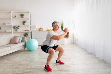 Morning training. Sporty mature man doing squats, exercising in living room interior, copy space