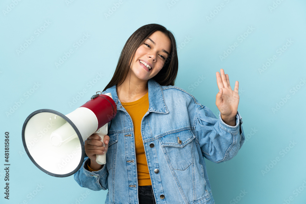 Wall mural teenager girl isolated on yellow background holding a megaphone and saluting with hand with happy ex