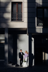 Fair-haired bride wearing wedding dress and lilac jacket and bridegroom in black suit standing at contrasting grey wall