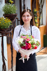Young woman in white shirt and apron holding bouquet of mixed unusual original flowers outdoors. Portrait of charming caucasian professional florist enjoying handmade, craft, working with flowers