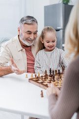 smiling senior man pointing with hand at chessboard near granddaughter and blurred wife.