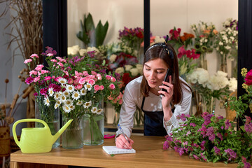 Female attractive florist accepting orders over phone and taking notes in notebook in flower shop....