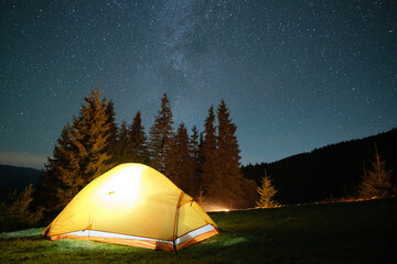 Bright illuminated tourist tent glowing on camping site in dark mountains under night sky with sparkling stars. Active lifestyle concept