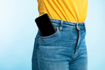 Unrecognizable woman having smartphone with empty screen in her jeans pocket on blue background,...