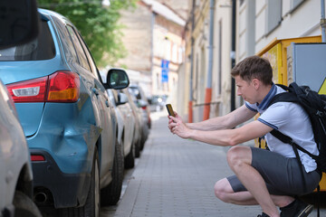 Stressed man driver taking photo on mobile phone camera of dented fender on street side for emergency service after car accident. Road safety and insurance concept