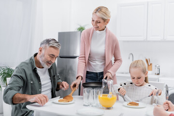 happy woman serving pancakes for husband during breakfast with grandchildren.