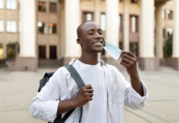 Happy black student guy taking off face mask, leaving university and walking in campus outdoors....
