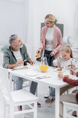 middle aged woman holding frying pan with pancakes near happy husband and grandchildren.